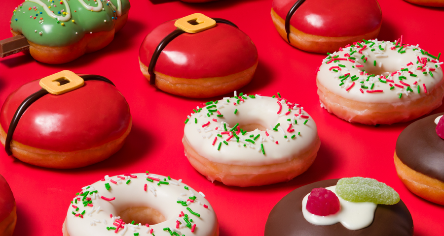 Krispy Kreme Christmas Doughnuts layed out on a red backdrop - featuring the Festive Sprinkles Ring Doughnut, the Santa Belly Filled Doughnut, the Christmas Pudding Doughnut and the Christmas Tree Shaped Doughnut