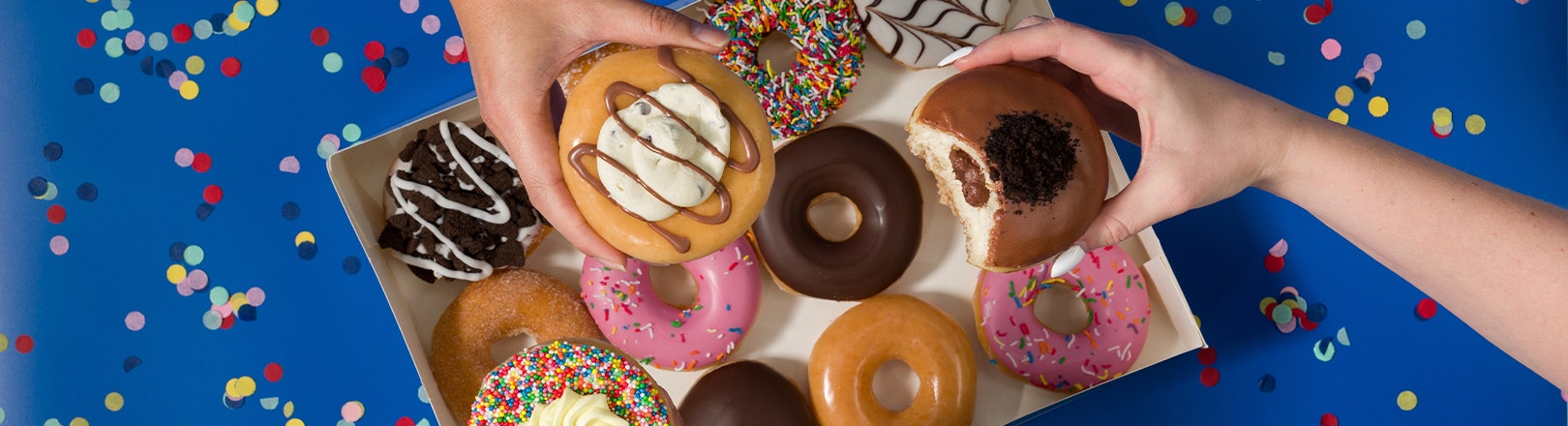 Krispy Kreme Limited Time Party Favourites Range Image of the party favourites dozen on a blue backdrop with a hand in frame holding one of the doughnuts in the range