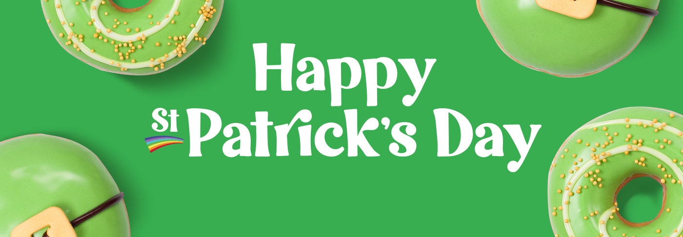 Krispy Kreme Limited Time Offer St Patrick's Day Doughuts Banner featuring the words Happy St Patrick's Dayu in white text on a green background. Surrounded by green iced doughnuts, one with a belt buckle decoration, the other with a white spiral and gold sprinkles